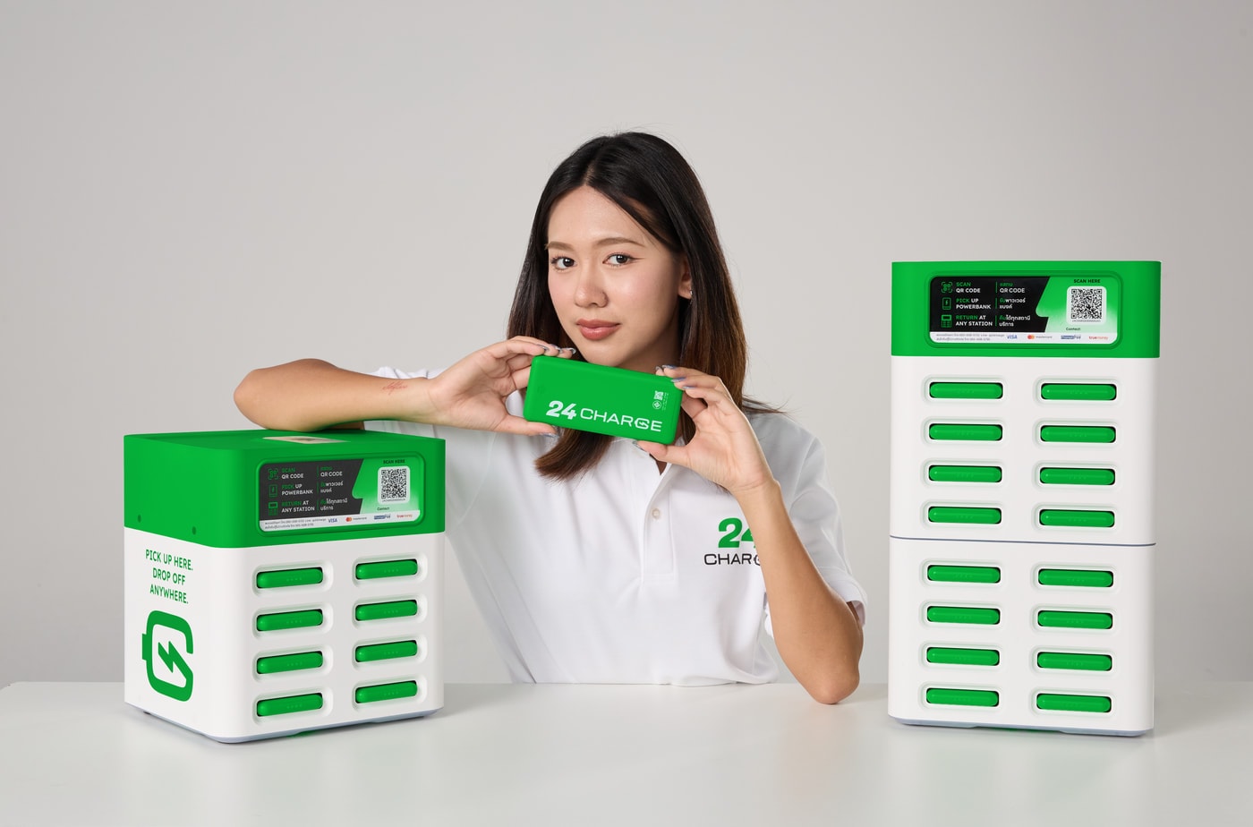 ผู้หญิงยืนถือพาวเวอร์แบงก์ 24CHARGE ระหว่างตู้เช่ารุ่น 8 ช่องและตู้ 16 ช่องที่วางอยู่บนโต๊ะ